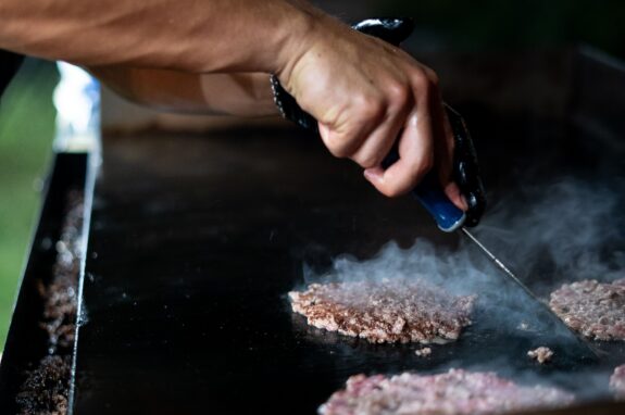 Hands using a spatula to cook beef patties on an outdoor flat-top griddle at night