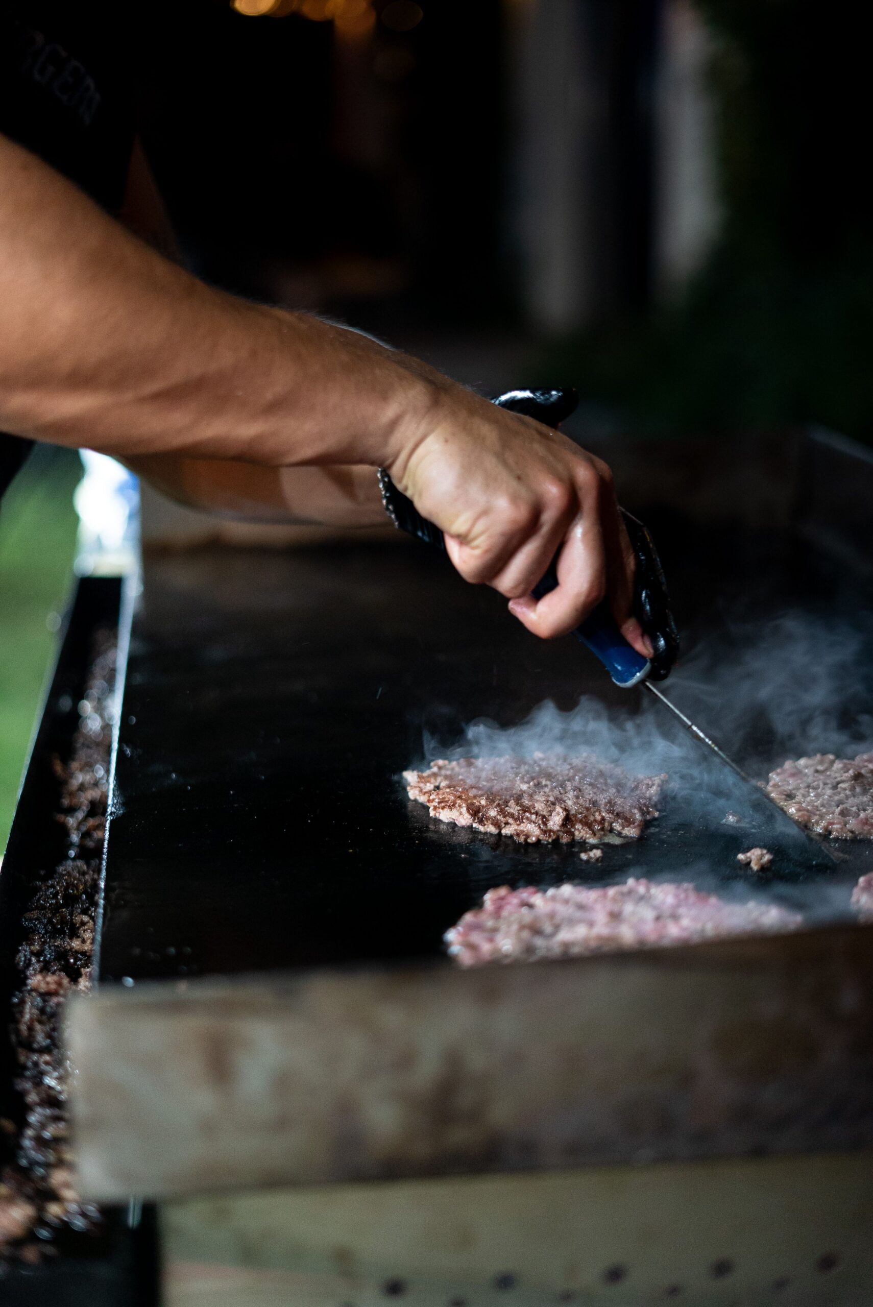 Hands using a spatula to cook beef patties on an outdoor flat-top griddle at night