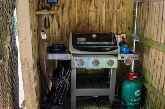 A gas grill with an attached propane tank set inside a wooden outdoor shelter.