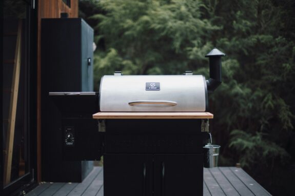 A black and silver outdoor pellet grill sitting on a backyard patio surrounded by lawn and landscaping.