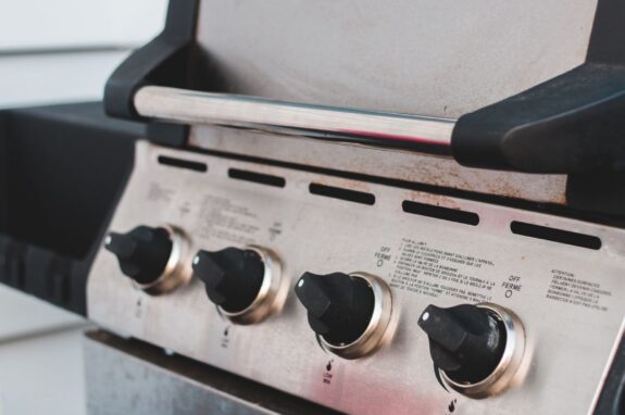 Close-up of a stainless steel gas grill with four black control knobs, a closed lid, and light rust specks along the vent slots