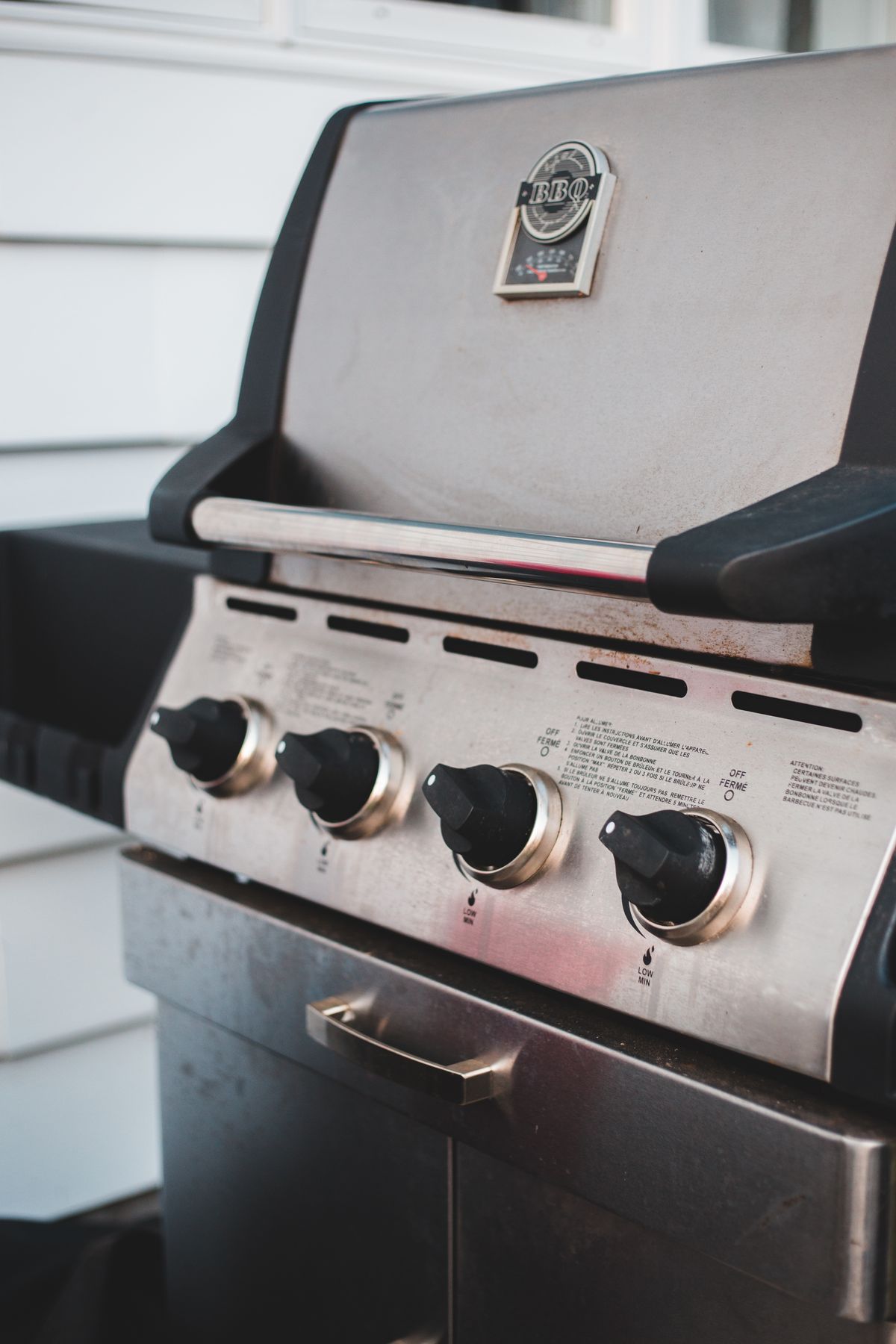 Close-up of a stainless steel gas grill with four black control knobs, a closed lid, and light rust specks along the vent slots